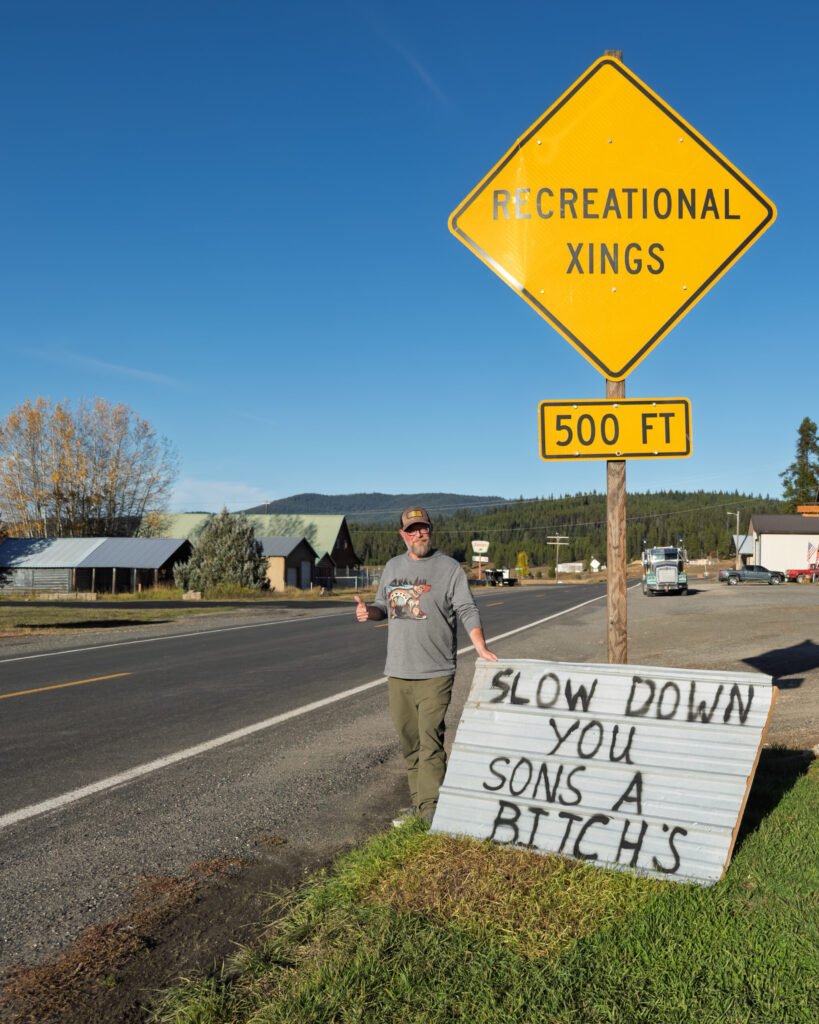 A hand-painted corrugated metal sign reading "slow down you sons a bitch's" beside a yellow RECREATIONAL XINGS 500 FT sign at the edge of Elk City, Idaho